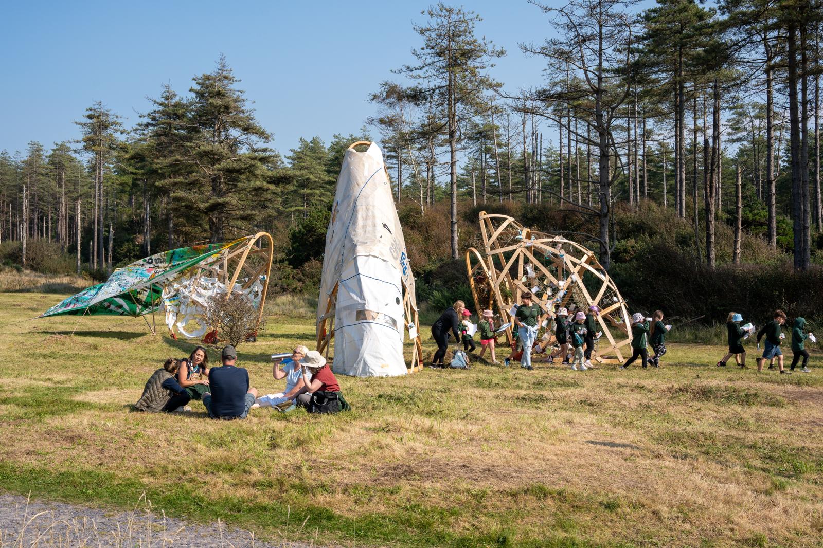 Large, temporary, bent wood, architectural structures sit arranged in a rough circular pattern on a grassy clearing next to a pine forest on a sunny day. The structures are constructed with lengths of wood fastened together in a mesh line pattern that forms an arch. They are organically decorated with sail cloths, string, and pieces of paper that have been attached by hand. A group of young children, all wearing green hoodies and with some wearing sun hats are walking out from between the structures holding pieces of paper looking like they’re off on a mission. They are joined by two adults. In the centre-left foreground, 5 adults sit in a circle looking like they’re having a discussion.