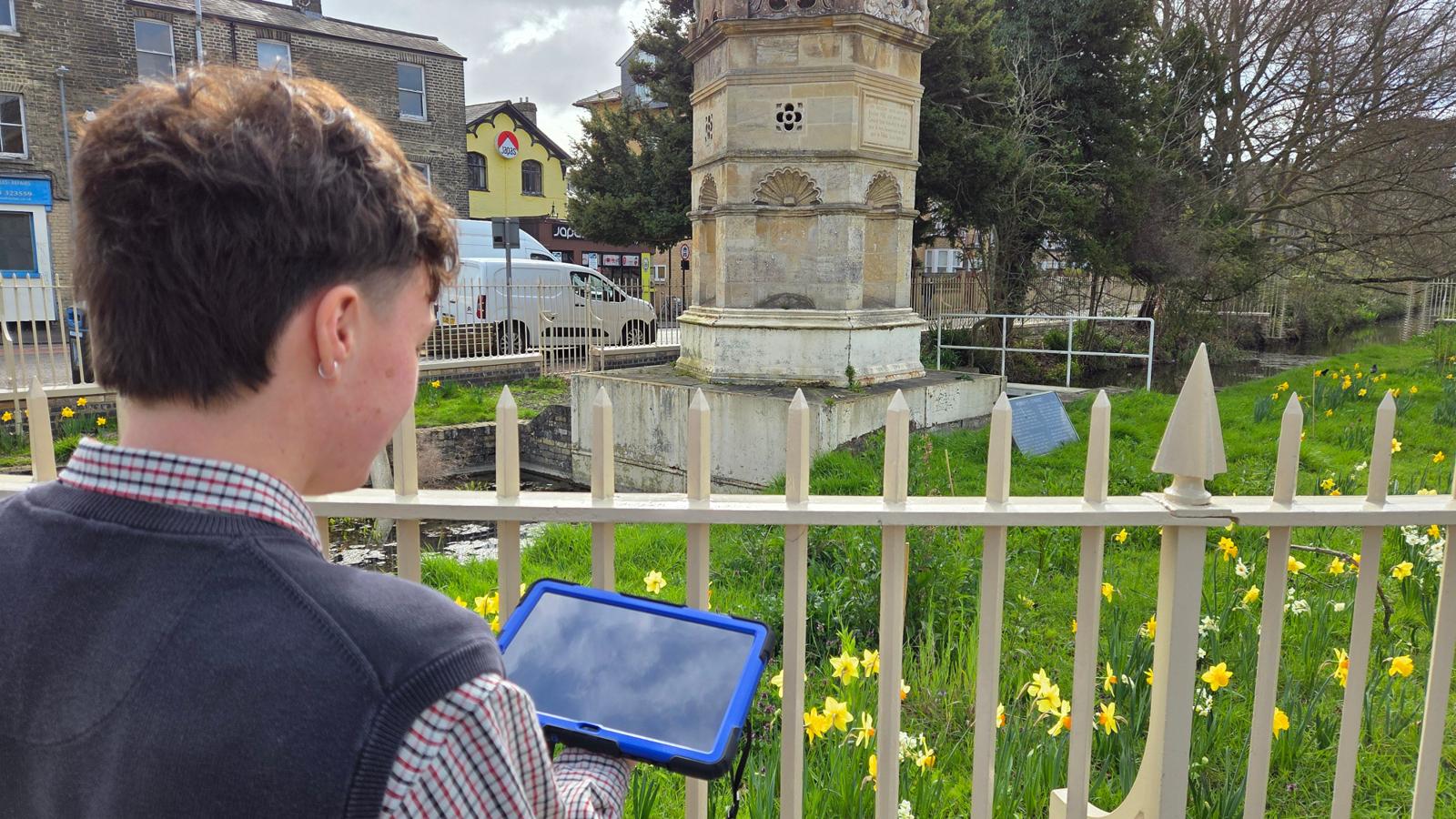 A person stands behind fence holding a blue‑cased tablet mapping, looking toward a historic Hobson's Conduit surrounded by green grass and blooming yellow daffodils.