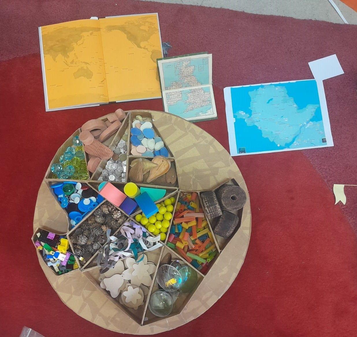 The Mandala of Ynys Môn: An outline model of Ynys Môn made of cardboard. The inside of it is divided into sections, each part filled with a different kind of object, e.g., corks, spectacle lenses, stones, LEGO, stones, pine cones, marbles and shoelaces.